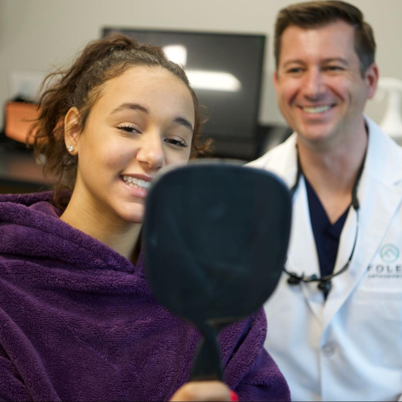 Young patient smiling while holding a mirror, showcasing her teeth, with Dr. Foley in a white coat, demonstrating a friendly orthodontic consultation experience at Foley Orthodontics in Cary, NC, emphasizing Invisalign and clear aligner treatment options.